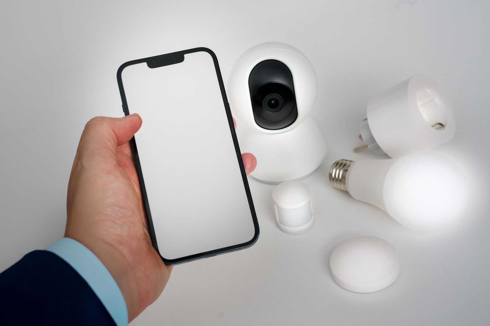 Main tenant un smartphone à côté d'objets connectés pour la maison : caméra, prise et ampoule
